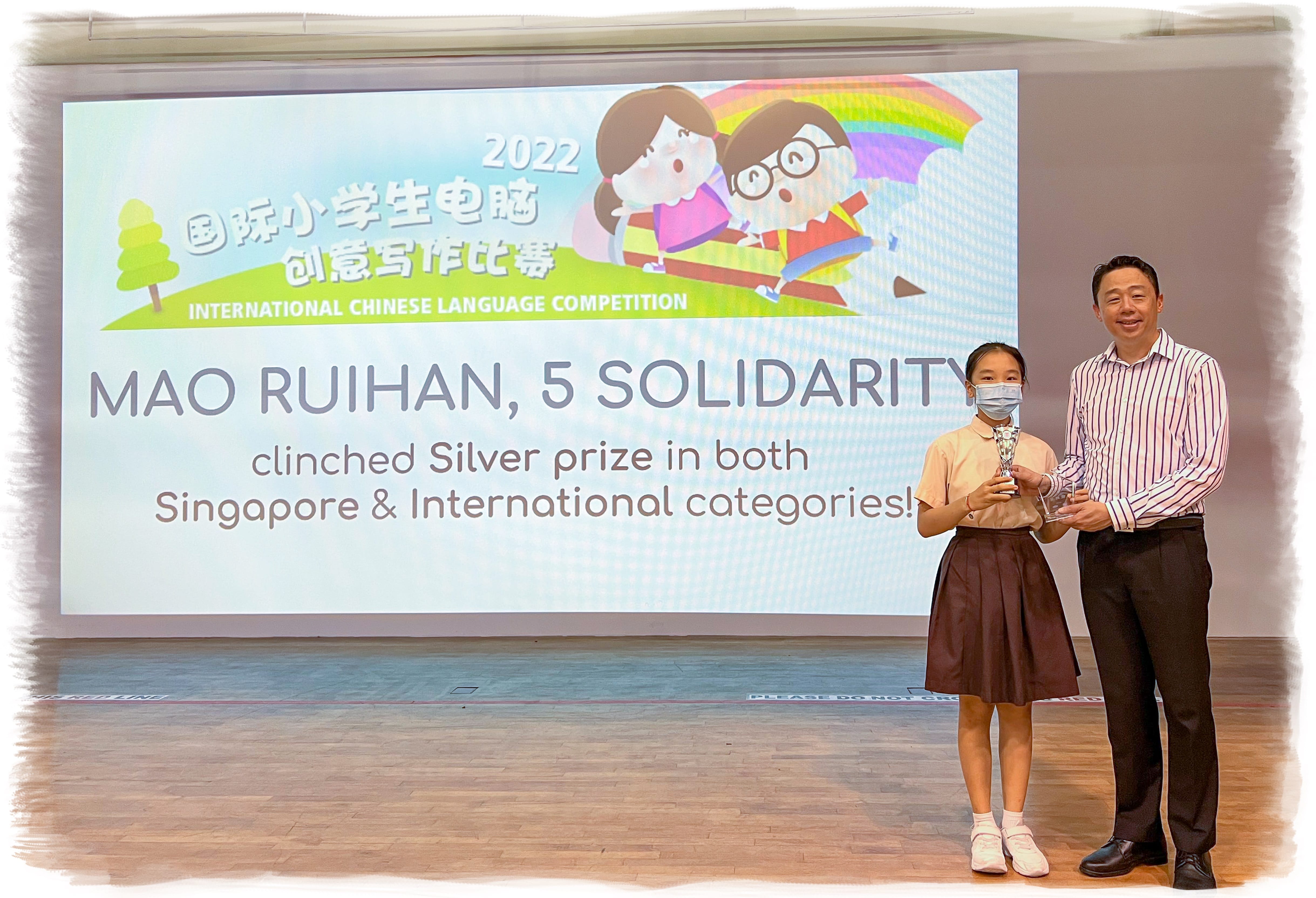 A girl and a man holding trophies stand before a screen announcing a 2022 International Chinese Language Competition Silver prize win.
